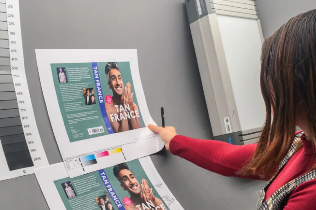 A person reviews printed book‑cover proofs featuring a large photo of Tan France and bold white text on a blue background. They point at the sheet with a pen while a grayscale colour calibration chart sits beside the prints, suggesting a design or print‑quality check in a studio workspace.