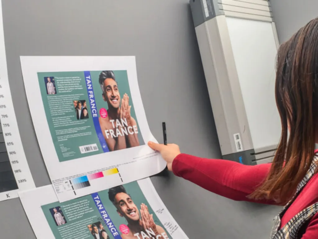 A person reviews printed book‑cover proofs featuring a large photo of Tan France and bold white text on a blue background. They point at the sheet with a pen while a grayscale colour calibration chart sits beside the prints, suggesting a design or print‑quality check in a studio workspace.