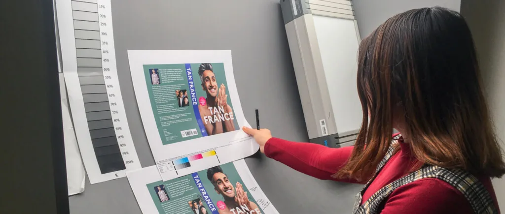 A person reviews printed book‑cover proofs featuring a large photo of Tan France and bold white text on a blue background. They point at the sheet with a pen while a grayscale colour calibration chart sits beside the prints, suggesting a design or print‑quality check in a studio workspace.