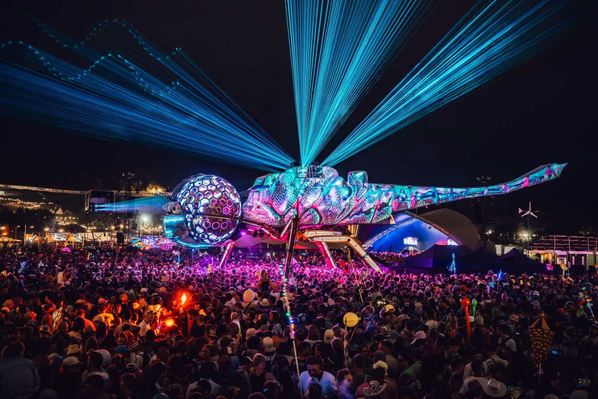 A giant biomechanical dragonfly stage shoots blue lasers over a massive festival crowd at night.