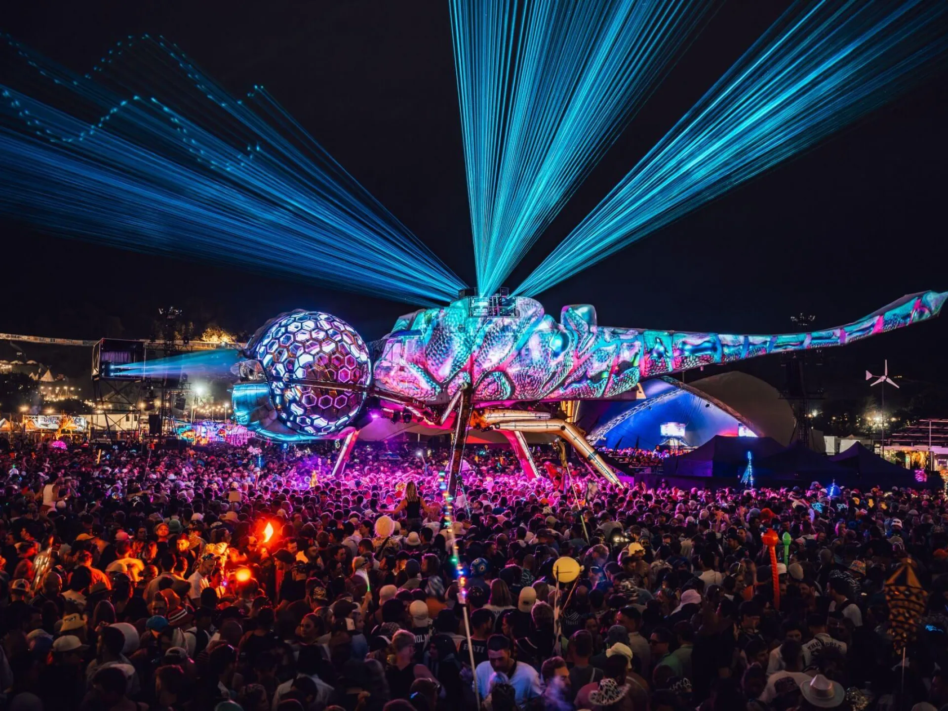 A giant biomechanical dragonfly stage shoots blue lasers over a massive festival crowd at night.