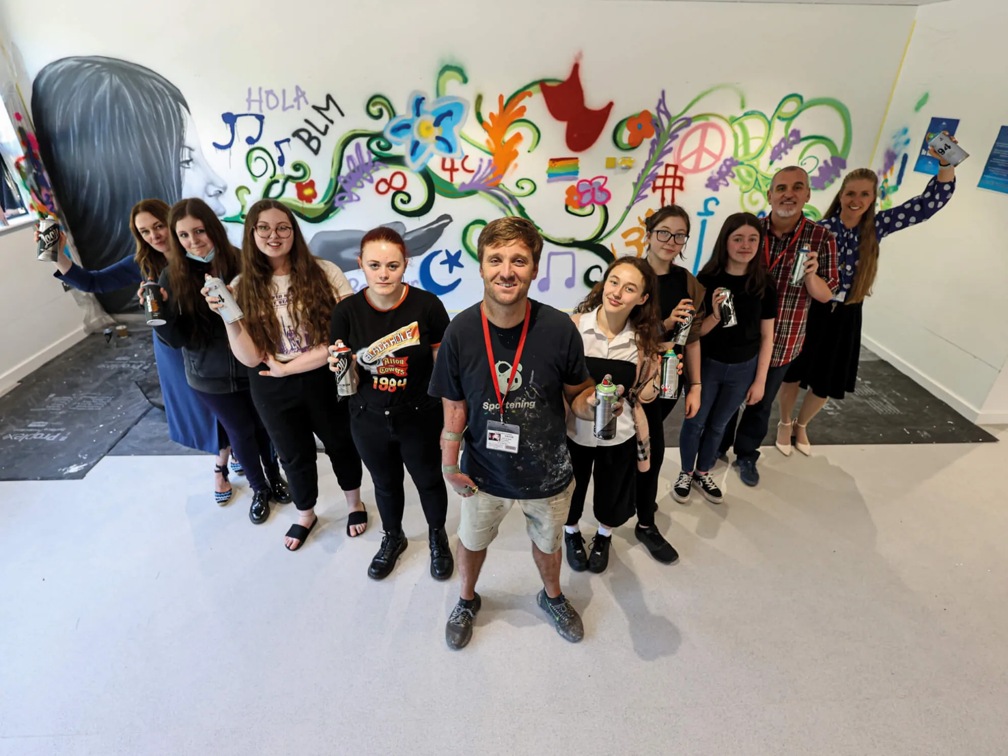 A group of people including teachers and young women, in a V-shape holding spray cans in front of graffiti art.
