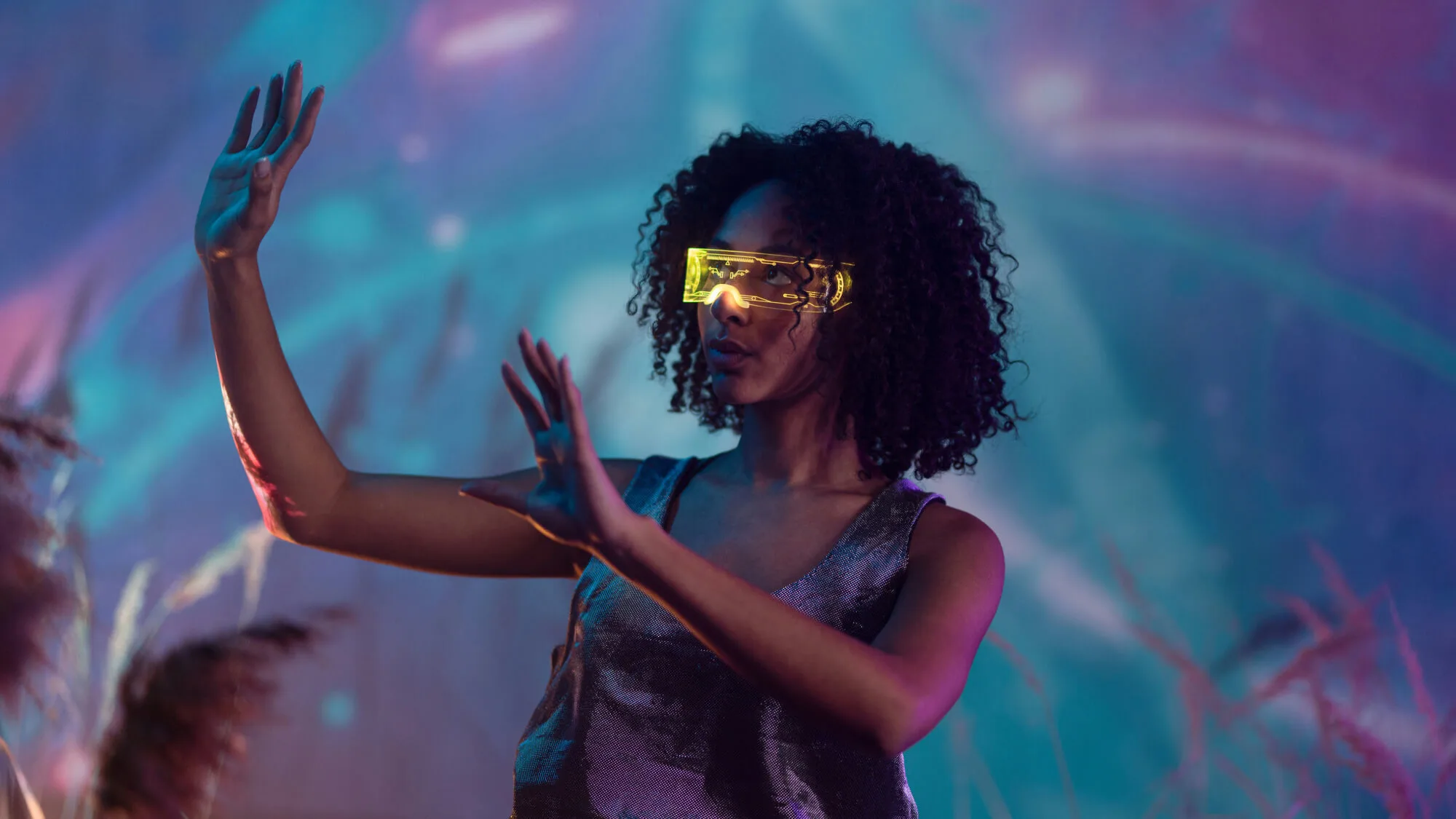 A woman with her hands raised to her right with interactive yellow goggles in front of a purple and blue space looking background.