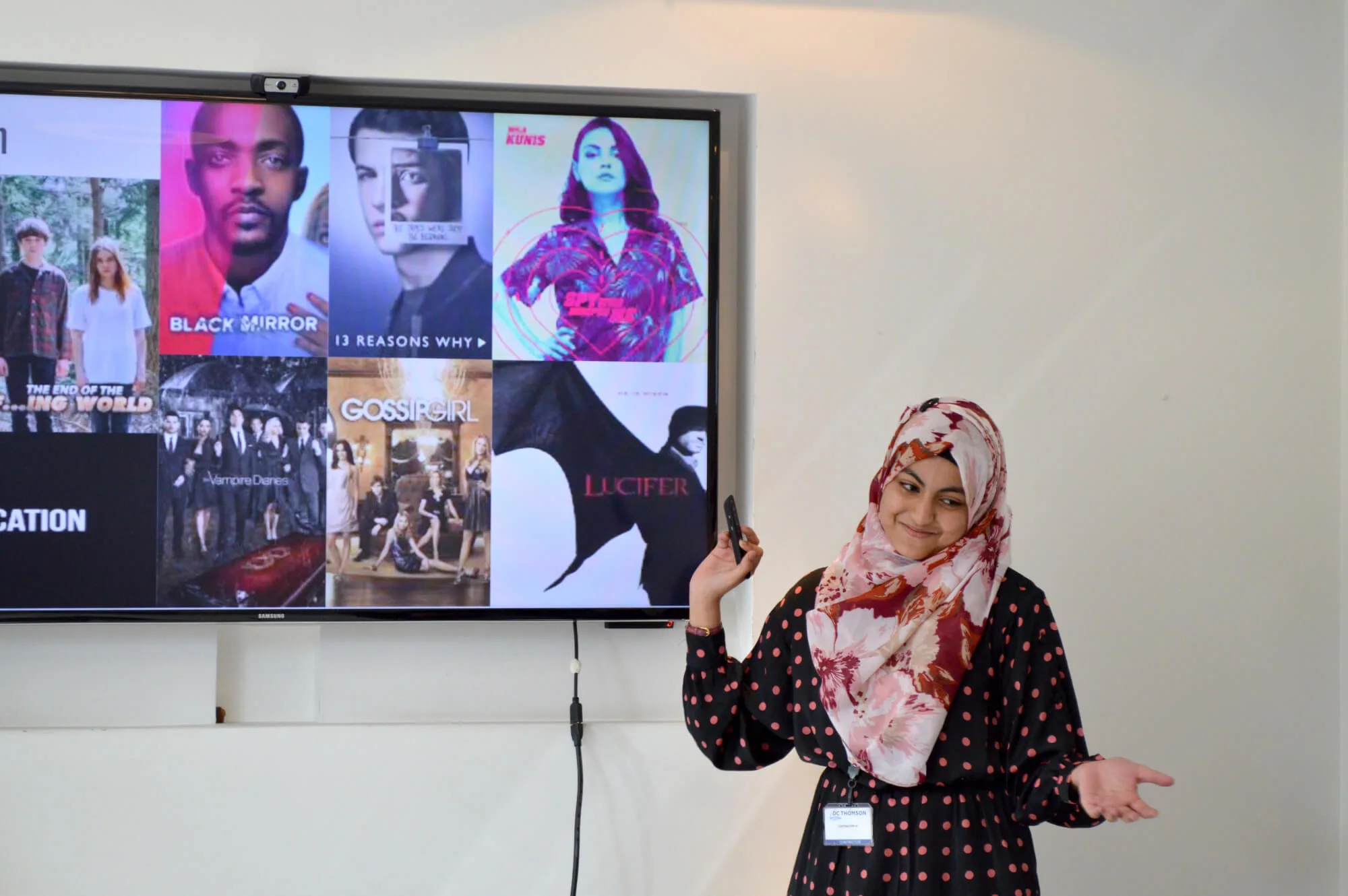 Woman pointing towards a screen showcasing TV shows including Lucifer, Black Mirror, 13 Reasons Why, The Vampire Diaries and Gossip Girl.