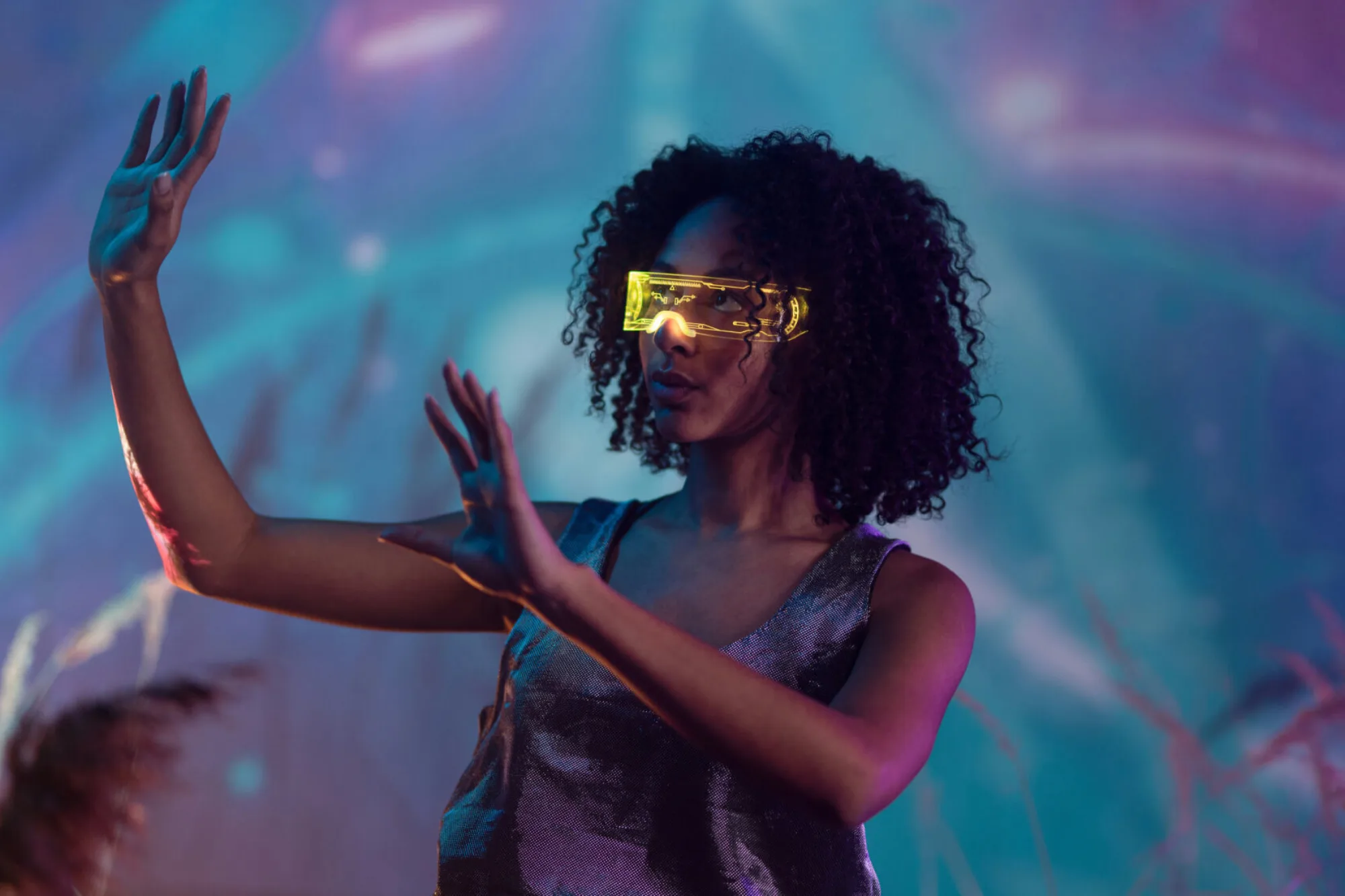 A woman with her hands raised to her right with interactive yellow goggles in front of a purple and blue space looking background.