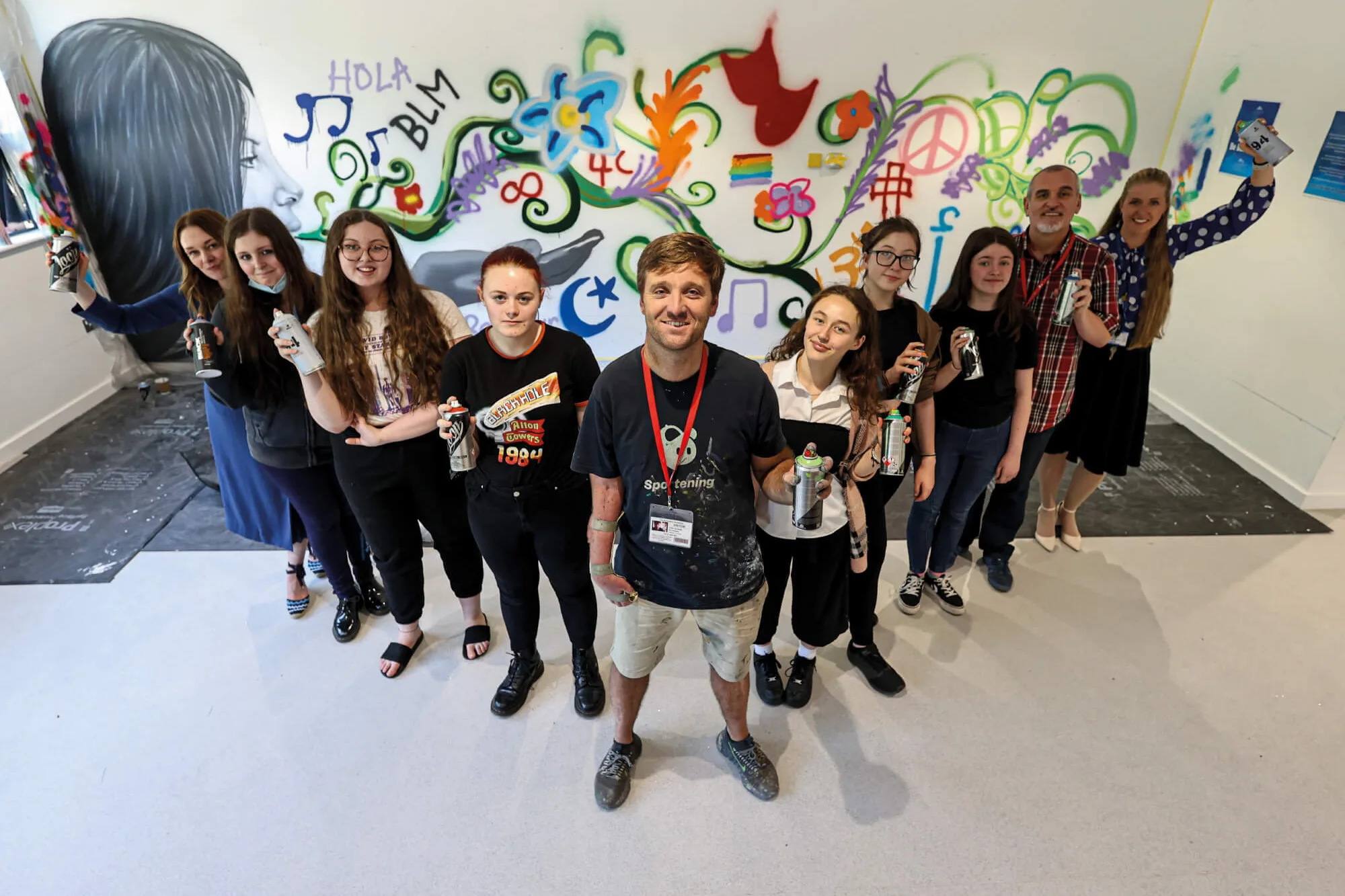 A group of people including teachers and young women, in a V-shape holding spray cans in front of graffiti art.