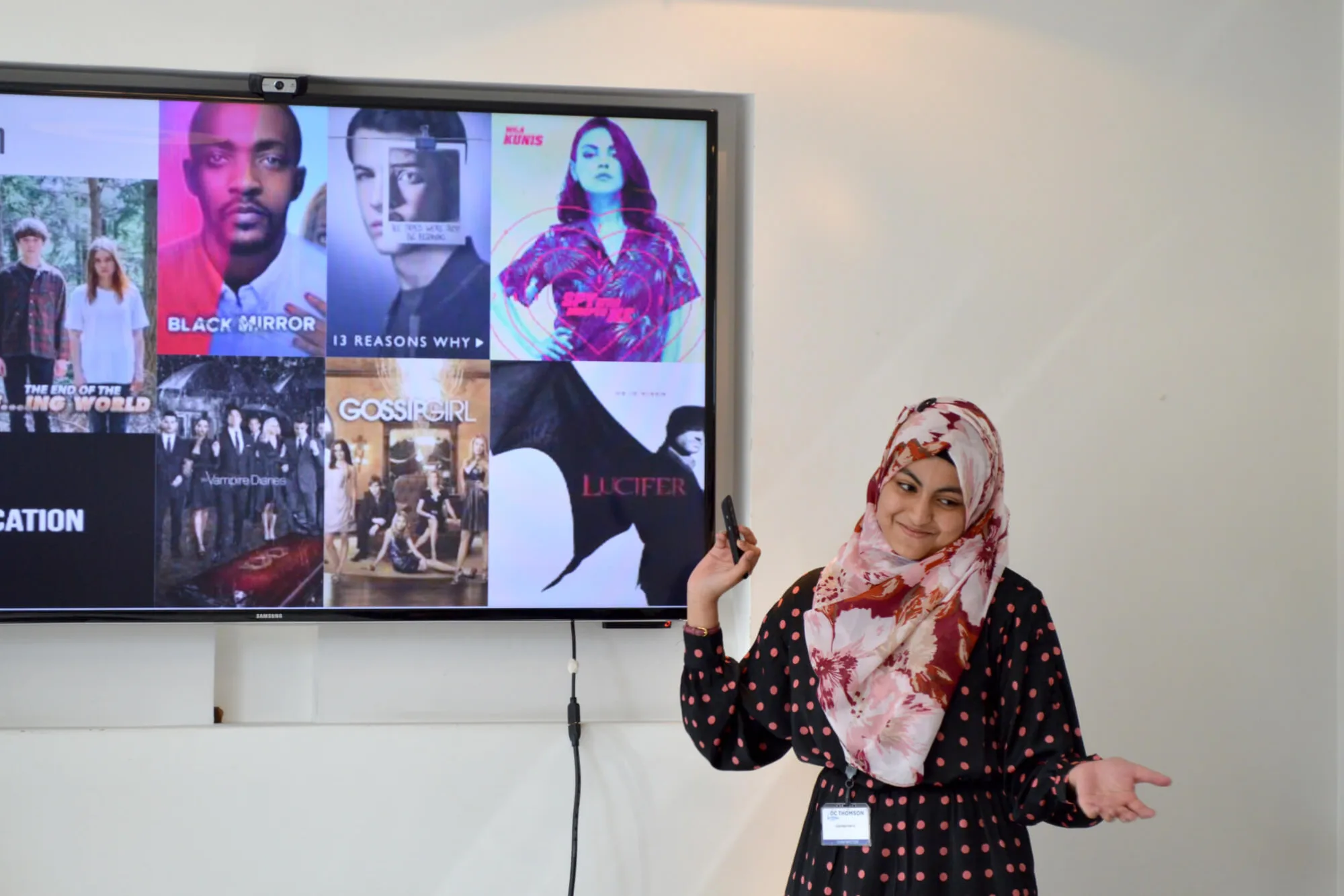 Woman pointing towards a screen showcasing TV shows including Lucifer, Black Mirror, 13 Reasons Why, The Vampire Diaries and Gossip Girl.