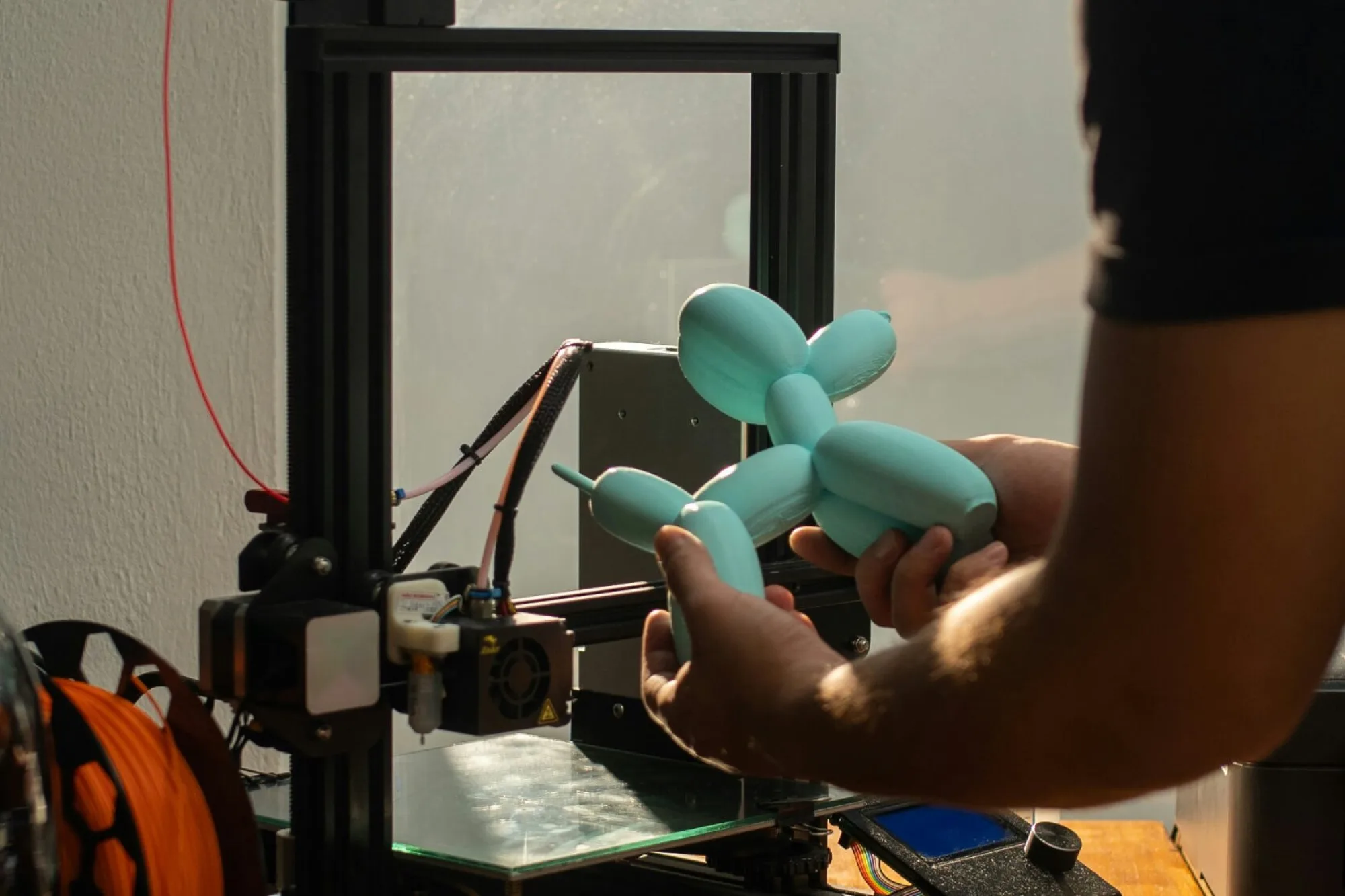 A person operates a 3D printer on a wooden table near a sunlit window, holding a light blue 3D‑printed balloon‑dog figure. The printer has a spool of red filament on top and an orange spool nearby, with the workspace lit by natural light.