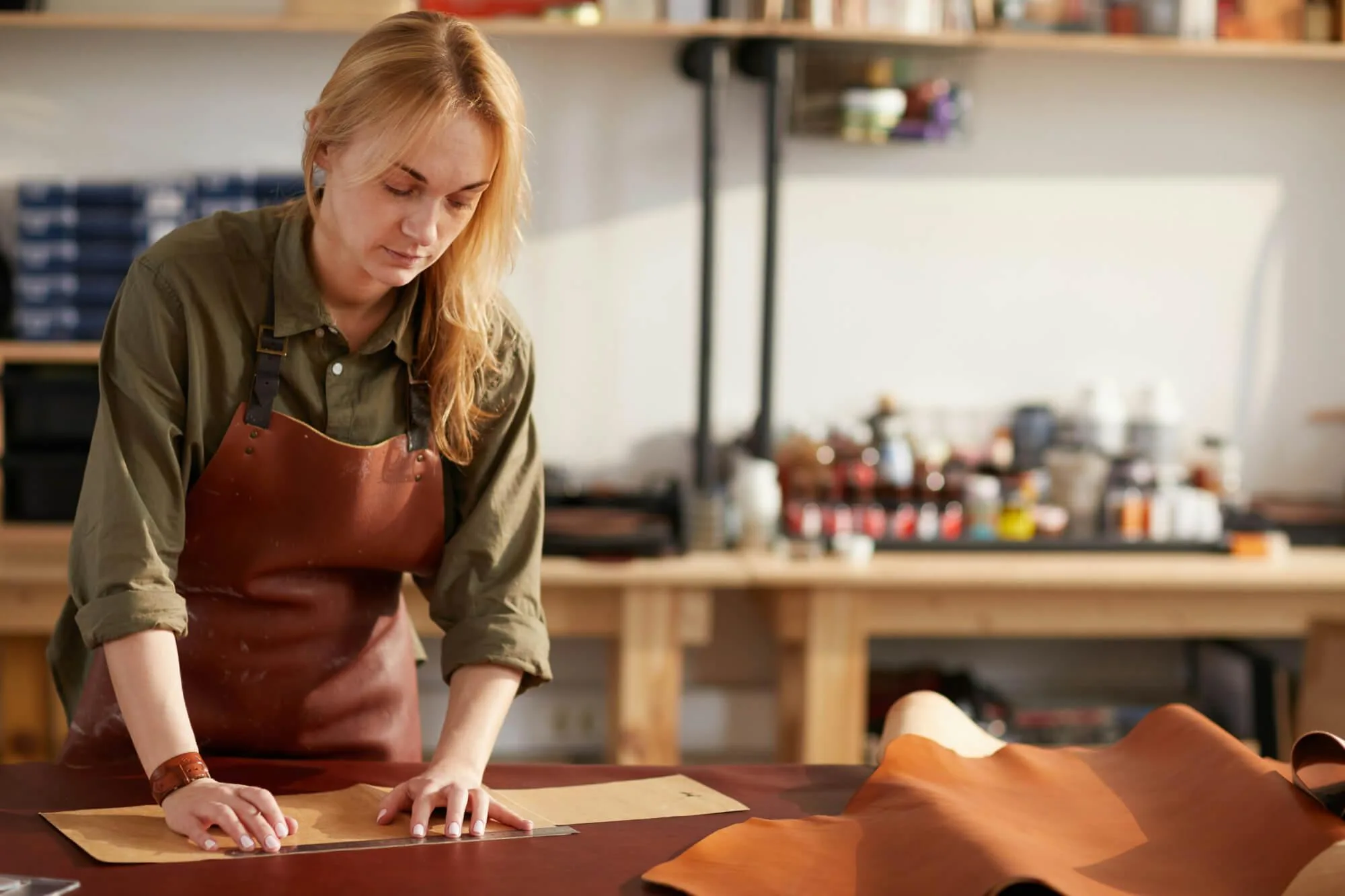 A person wearing a brown leather apron works at a studio bench, cutting pieces of leather with a hand tool. Shelves in the background hold tools, materials, and containers, creating a focused leather‑crafting workspace.