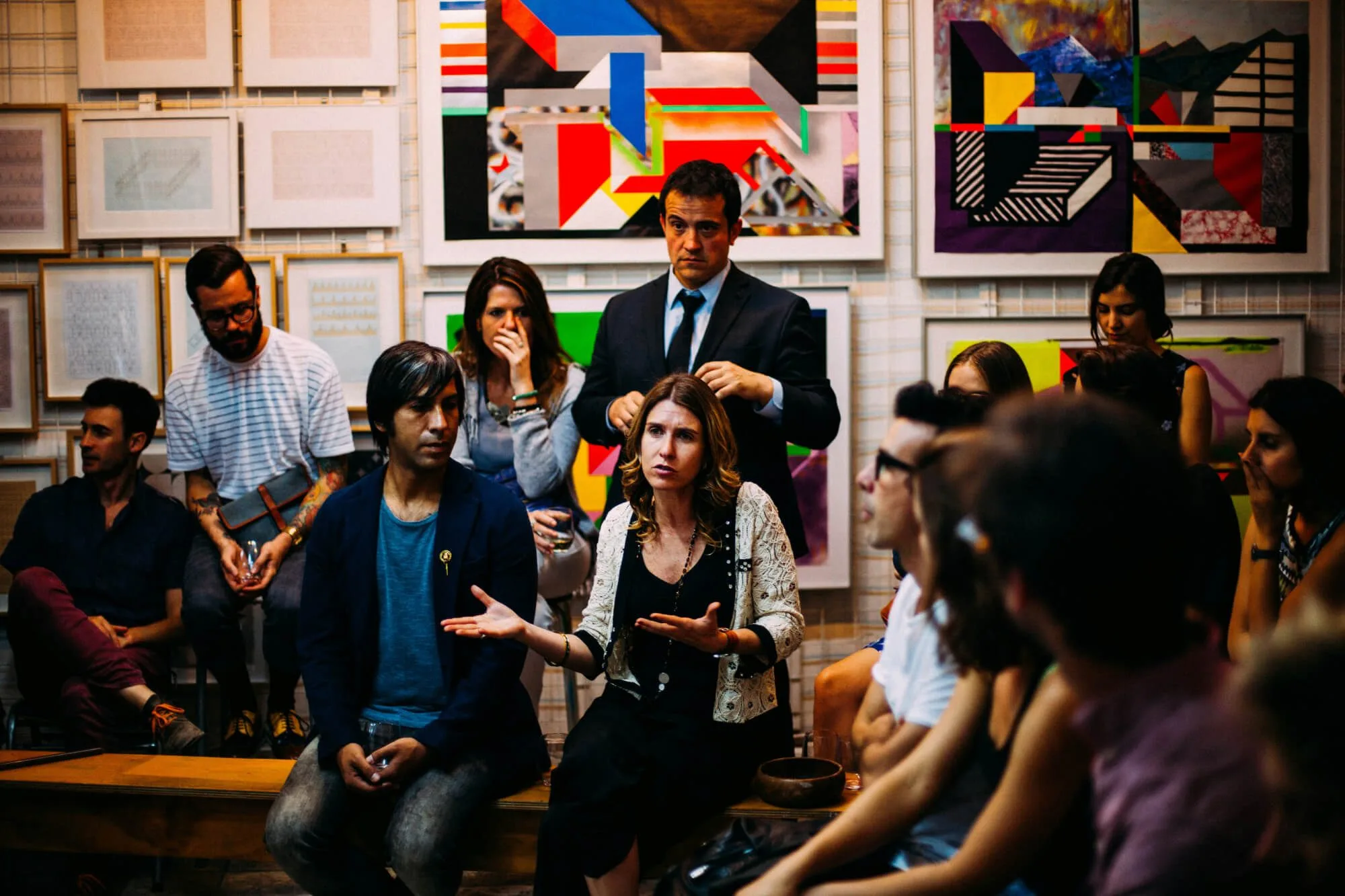 A group of people gather in an art‑filled room, seated and standing as they engage in a lively discussion. One person gestures while speaking, and colourful framed artworks cover the wall behind them, creating a creative, conversational atmosphere.