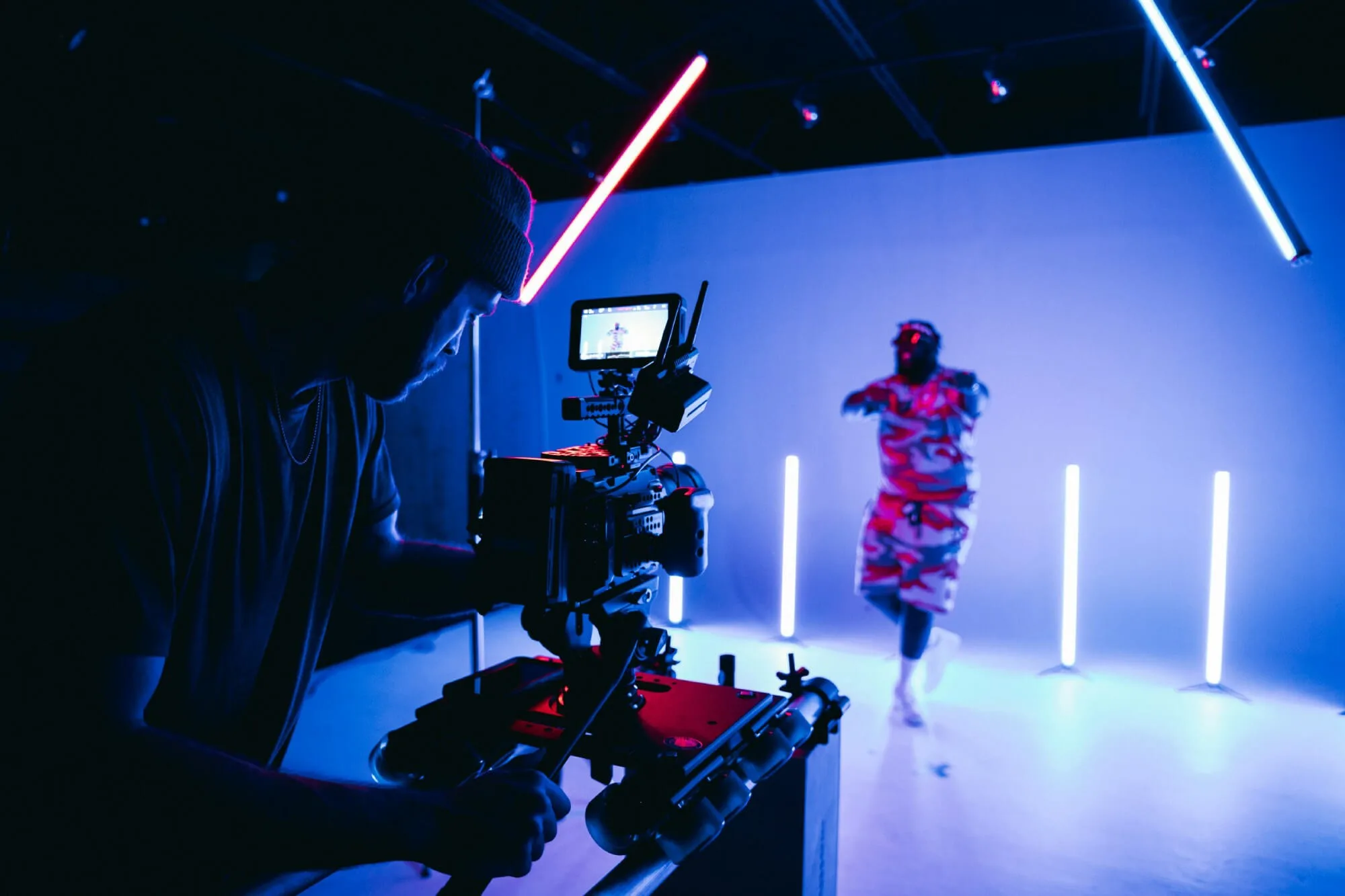 A camera operator films a performer on a blue‑lit set, using a professional camera mounted on a stabilizing rig. The monitor shows the subject being recorded, while bright pink and blue LED light tubes create a futuristic backdrop around the scene.