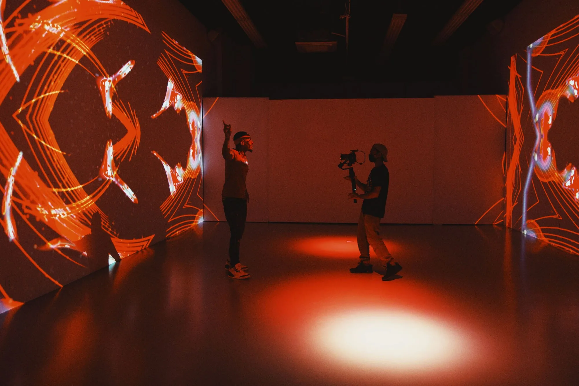 Two people work in a dark room lit by vivid red and orange projected patterns. One person stands and gestures toward the glowing geometric visuals on the walls, while the other films with a camera on a stabilizer. The reflective floor and immersive lighting create a dramatic, cinematic atmosphere.