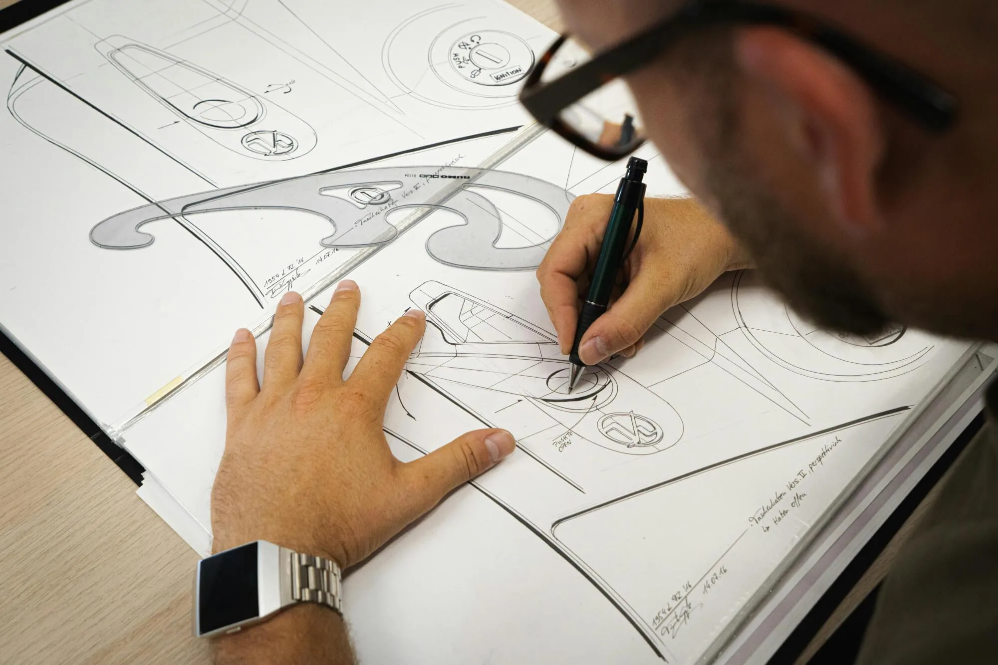 A person sketches detailed industrial design concepts on large sheets of paper, using a pen and a French curve template to create smooth, precise lines. Multiple aerodynamic product drawings cover the workspace, and the person wears a metallic wristwatch and glasses while working.