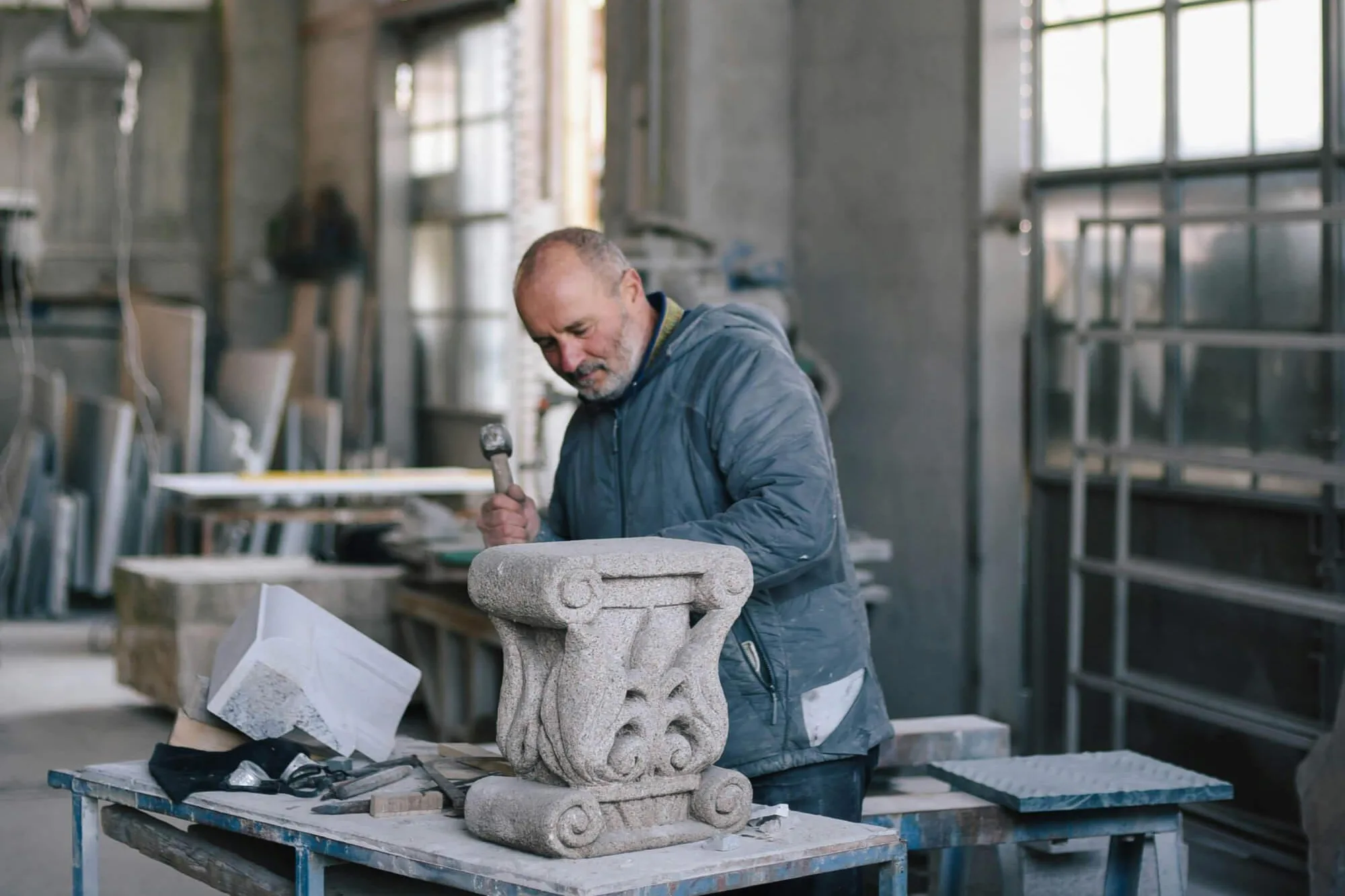 A person works in a bright workshop, carving a decorative stone piece with scroll and leaf details. Tools, wooden planks, and stone fragments sit on the table around them, and large windows illuminate the industrial studio space.
