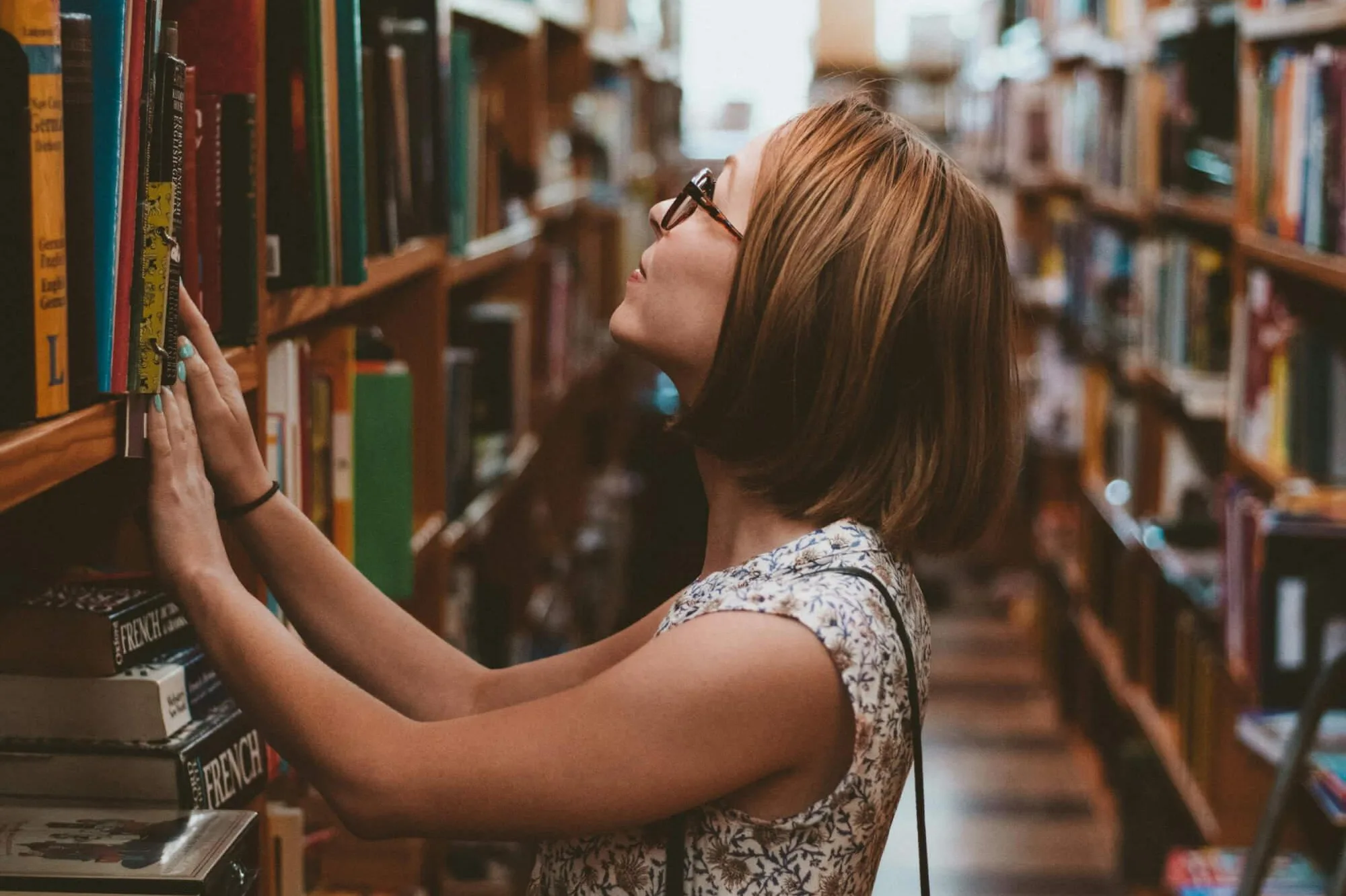 A person stands between tall wooden bookshelves in a warm, softly lit library or bookstore, reaching toward the shelves while wearing glasses and a light floral outfit. Rows of books surround them, and a checkered floor leads toward a bright area in the background.