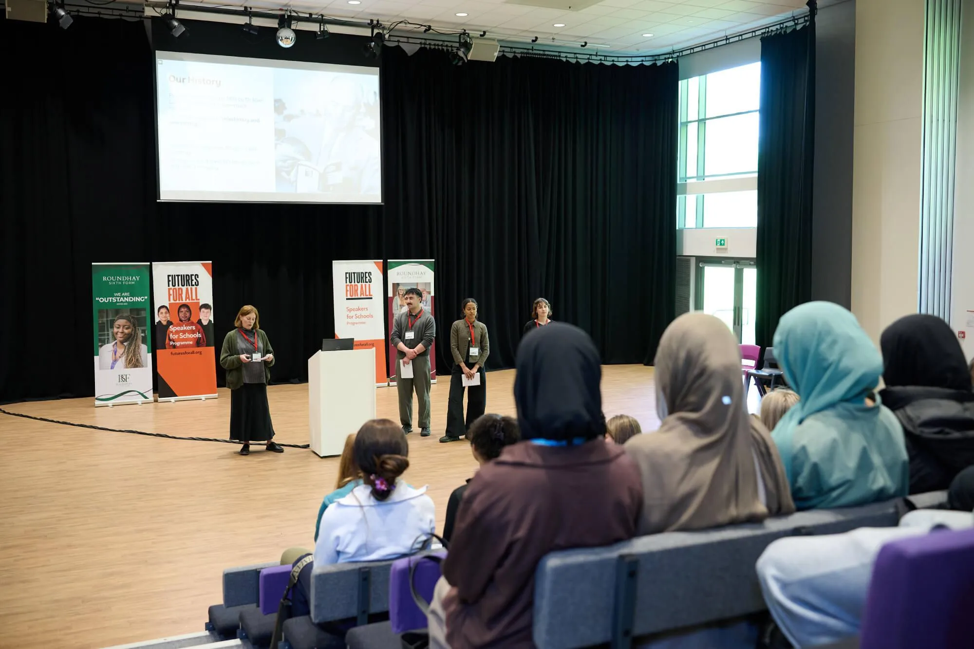 Four presenters stand at the front of a school auditorium, speaking to students seated in tiered rows. Behind them, banners read “ROUNDHAY We are ‘OUTSTANDING’,” “FUTURES FOR ALL,” and “Speakers for Schools,” and a large screen displays a slide titled “Our History” with a black‑and‑white photo and text.