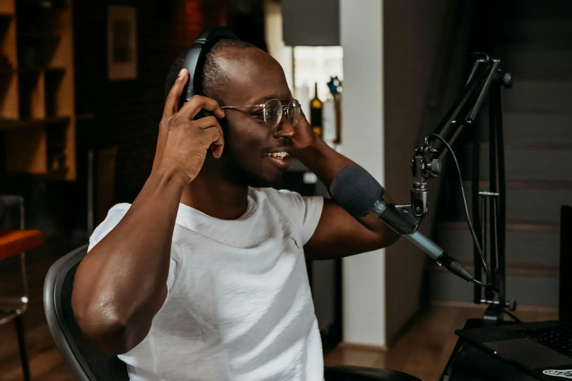 A person wearing headphones speaks into a mounted microphone at a desk with a laptop, keyboard, and mouse. The modern indoor space includes a staircase, shelves with bottles, and framed artwork, creating a cozy recording or podcasting setup.