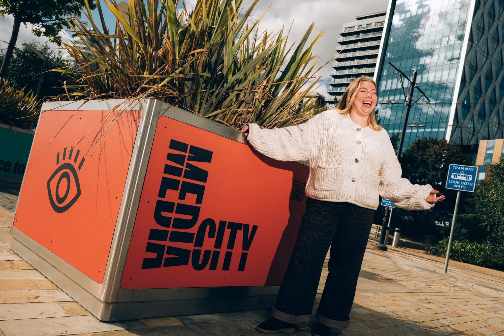 A person stands beside a large orange planter marked “MEDIA CITY” in bold black lettering, in an urban plaza surrounded by modern glass office buildings. Tall ornamental plants fill the planter, and tramway signage and paved walkways are visible in the background. The person is leaning against the planter and holding their arms out while standing in the foreground.