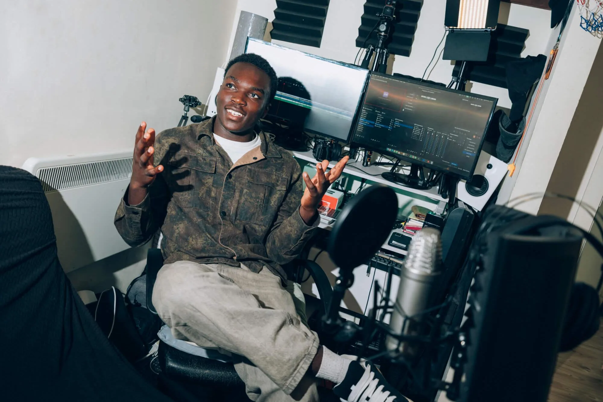 A person sits in a desk chair inside a compact home studio or content creation workspace, gesturing with both hands while facing someone off-camera. In the foreground, a large microphone with a pop filter is positioned on a boom arm, suggesting a podcast, recording, or streaming setup. Behind the person are two computer monitors displaying video editing software, along with a camera, studio lighting panels, acoustic foam panels, speakers, and other audio-visual equipment. The desk contains various accessories and electronic devices. The room is brightly lit, with white walls and a wooden floor, creating the appearance of a personal media production or editing studio.