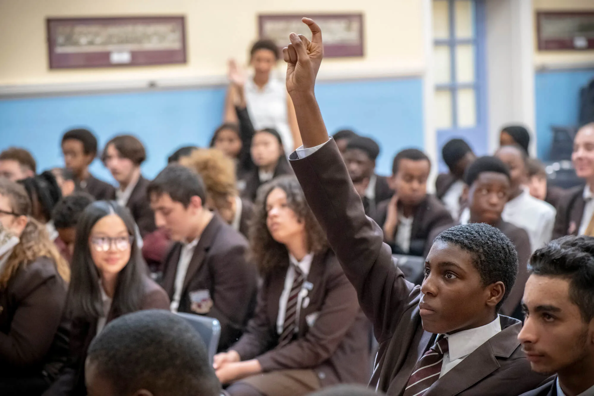Group of school students in an assembly focussed on one young man with his hand raised.
