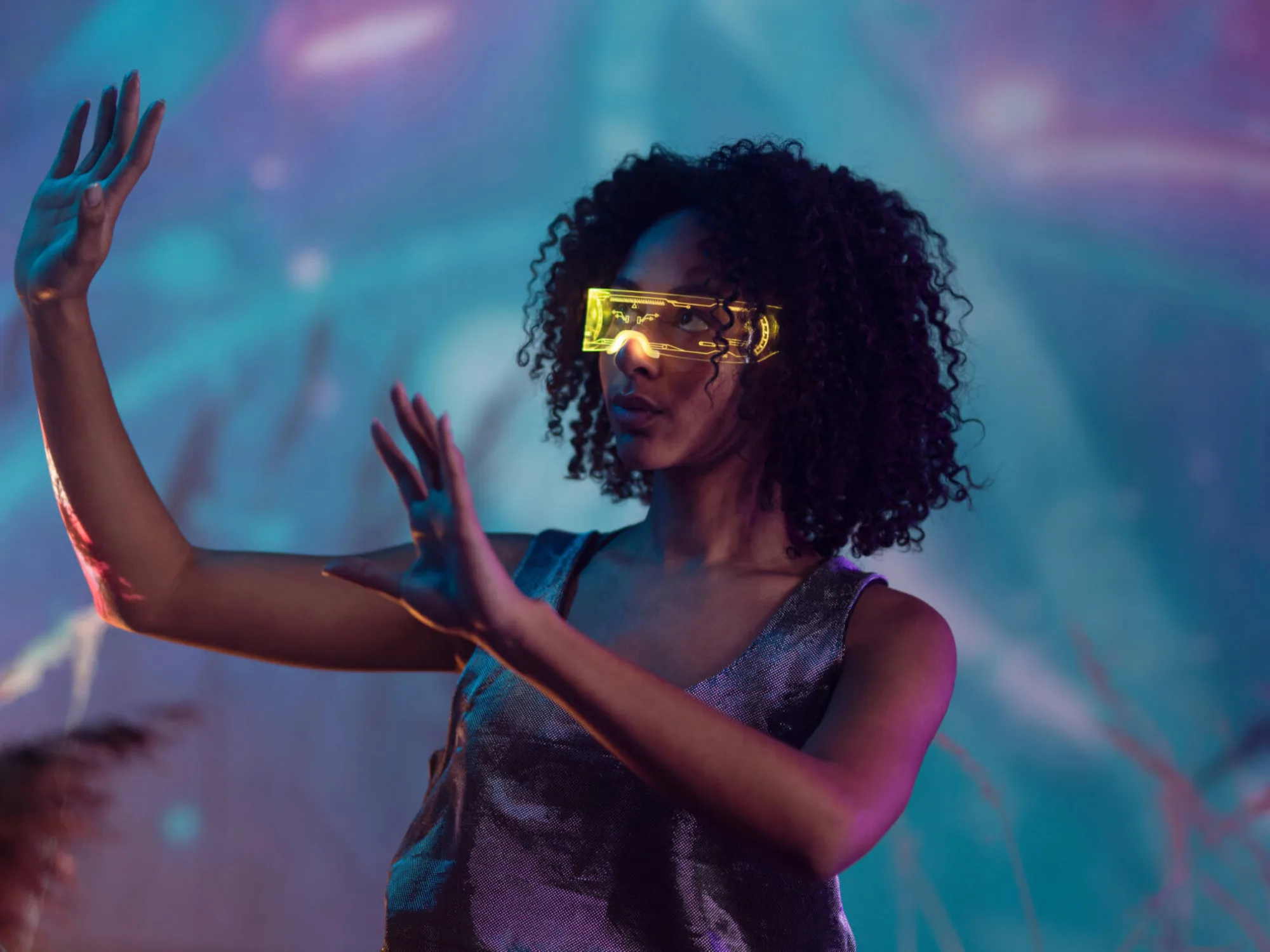 A woman with her hands raised to her right with interactive yellow goggles in front of a purple and blue space looking background.