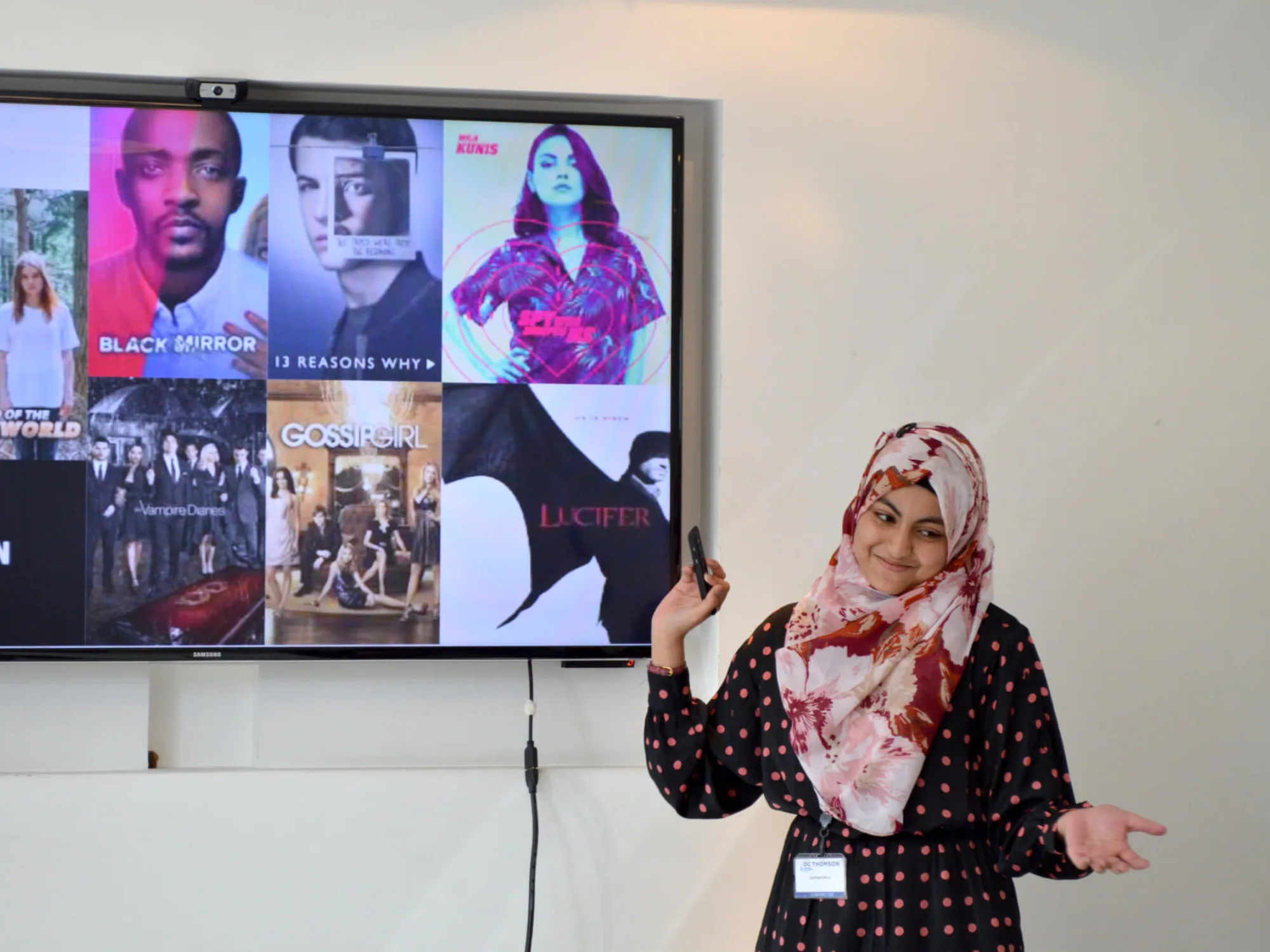 Woman pointing towards a screen showcasing TV shows including Lucifer, Black Mirror, 13 Reasons Why, The Vampire Diaries and Gossip Girl.