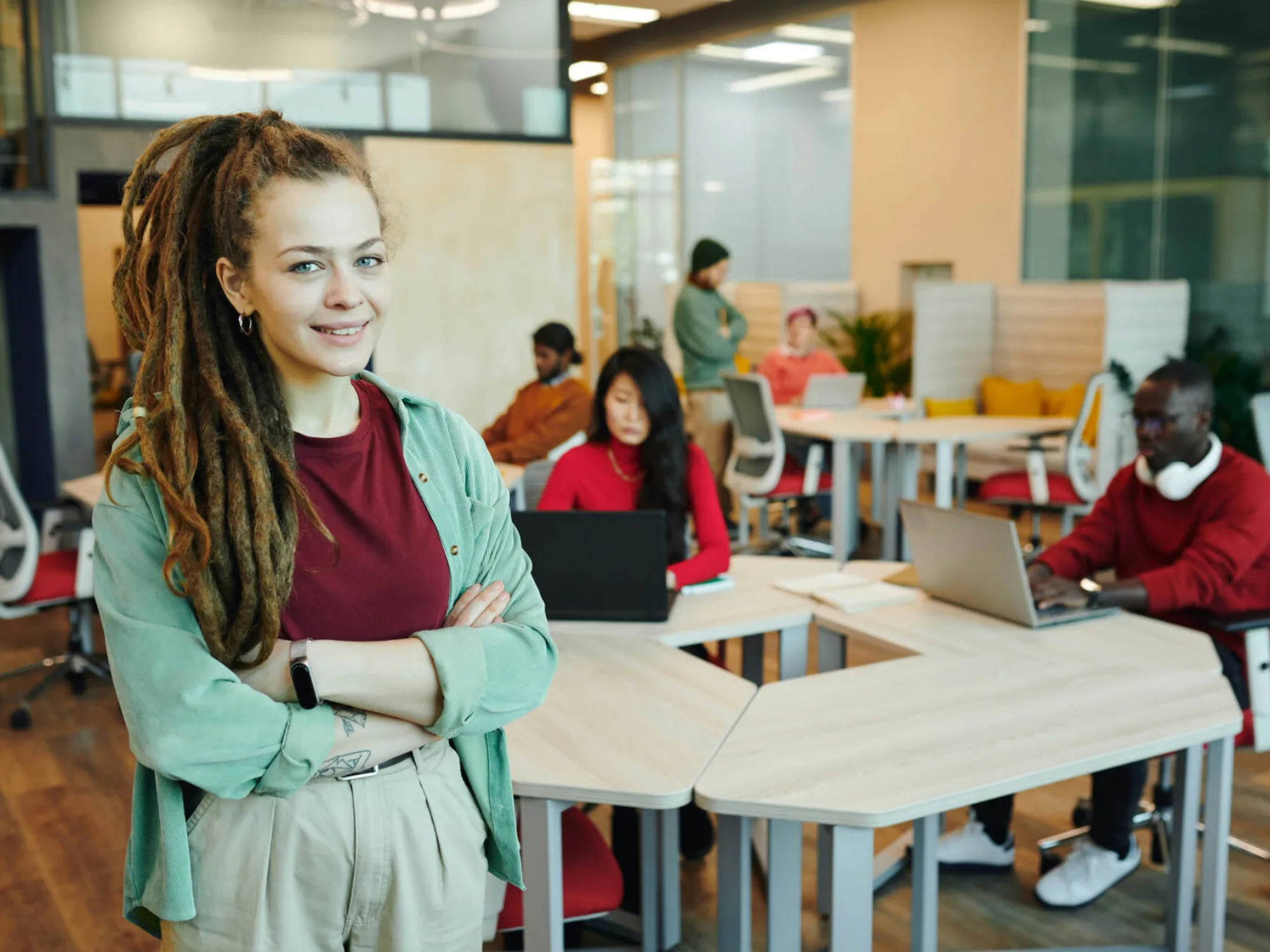 A woman smiling, standing with her arms folded at the front of an office space with men and women hot desking on their laptops.