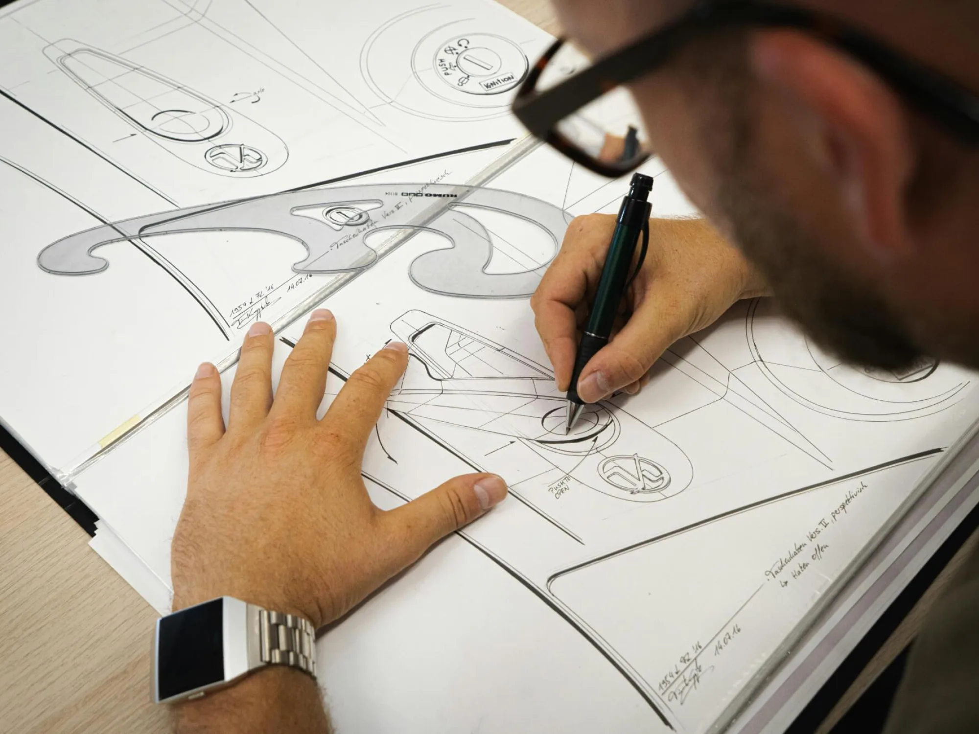 A person sketches detailed industrial design concepts on large sheets of paper, using a pen and a French curve template to create smooth, precise lines. Multiple aerodynamic product drawings cover the workspace, and the person wears a metallic wristwatch and glasses while working.