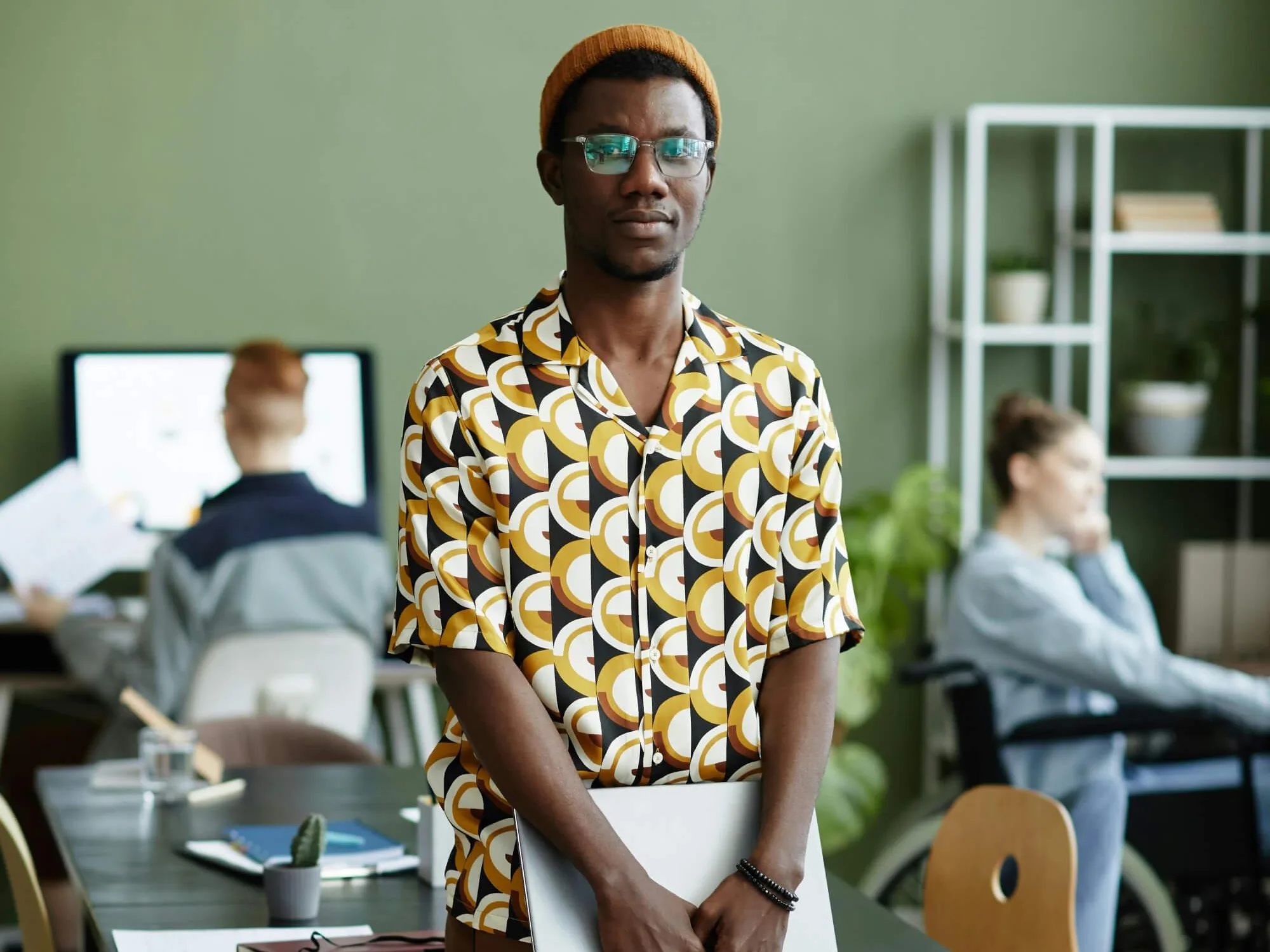 A person wearing a patterned shirt, brown trousers, tan shoes, and a mustard beanie stands in a modern office holding a closed laptop. Behind them, two coworkers sit at desks—one at a computer and one in a wheelchair. The workspace features a green wall, shelves with books and decor, and a black table with chairs.