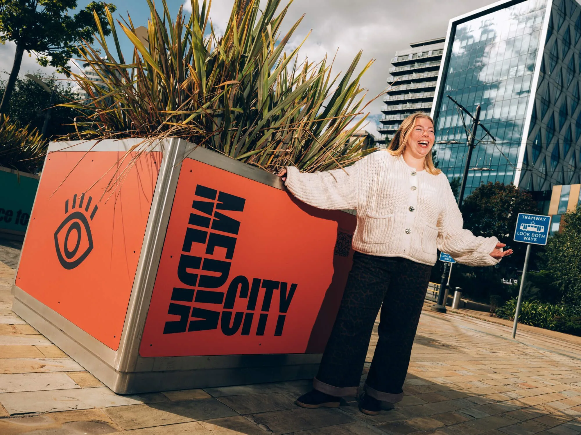 A person stands beside a large orange planter marked “MEDIA CITY” in bold black lettering, in an urban plaza surrounded by modern glass office buildings. Tall ornamental plants fill the planter, and tramway signage and paved walkways are visible in the background. The person is leaning against the planter and holding their arms out while standing in the foreground.