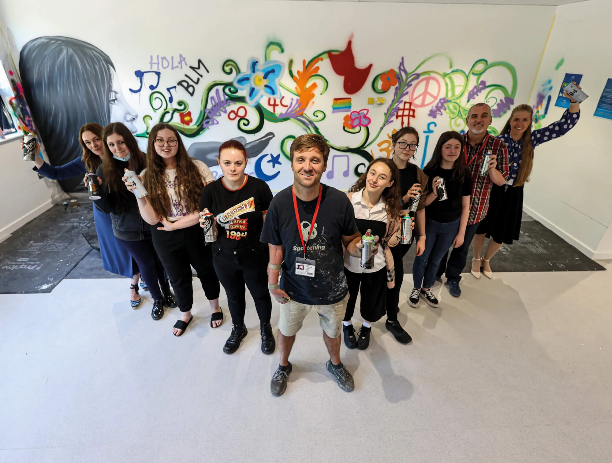 A group of people including teachers and young women, in a V-shape holding spray cans in front of graffiti art.