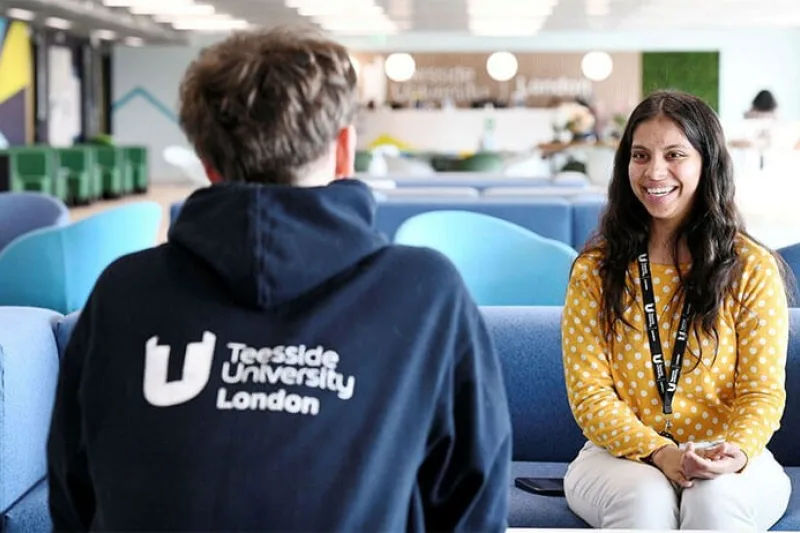 Two people sit facing each other on blue sofas in a modern university common area. In the foreground, one person is seen from behind wearing a navy hoodie with the text “Teesside University London” printed in white on the back. Opposite, another person wearing a yellow polka-dot top and a lanyard sits with hands clasped, engaged in conversation. The open-plan space features blue seating, bright overhead lighting, and contemporary décor, with additional seating and a café-style area visible in the background. The image conveys a student support, mentoring, or campus discussion setting within a university environment.
