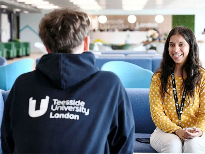 Two people sit facing each other on blue sofas in a modern university common area. In the foreground, one person is seen from behind wearing a navy hoodie with the text “Teesside University London” printed in white on the back. Opposite, another person wearing a yellow polka-dot top and a lanyard sits with hands clasped, engaged in conversation. The open-plan space features blue seating, bright overhead lighting, and contemporary décor, with additional seating and a café-style area visible in the background. The image conveys a student support, mentoring, or campus discussion setting within a university environment.